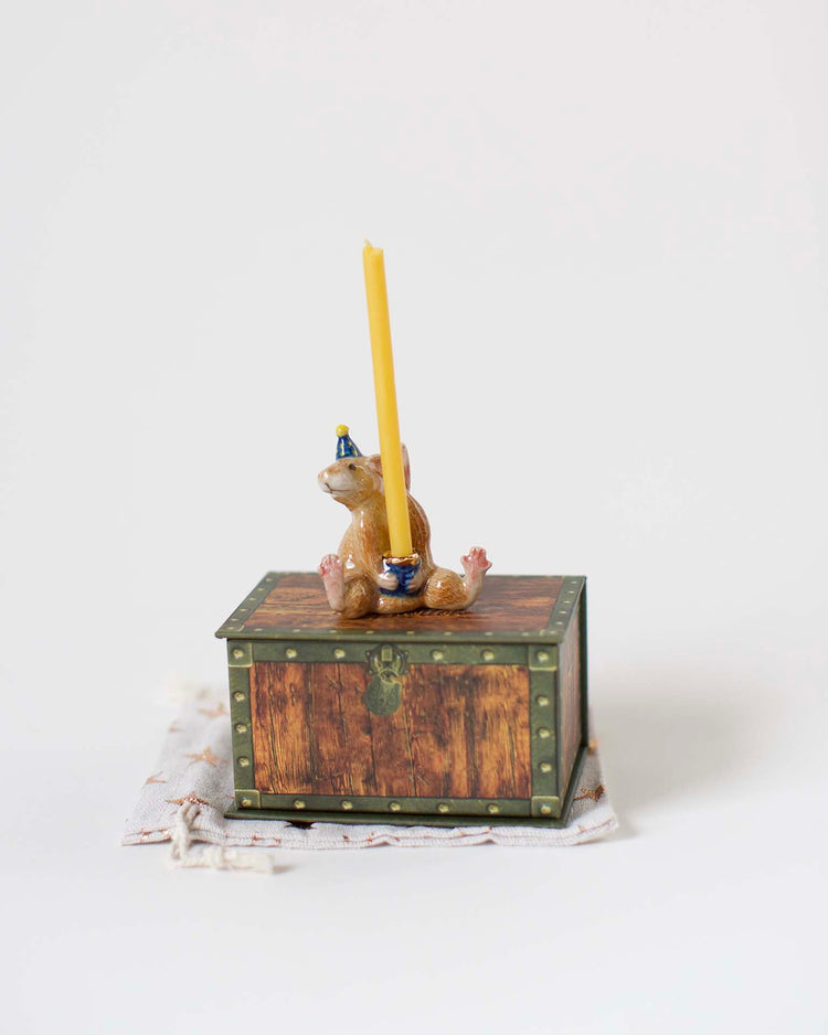 A hand-painted figurine of a year of the rat cake topper from Camp Hollow, sitting on a treasure chest and holding a yellow candle, set against a plain white background.