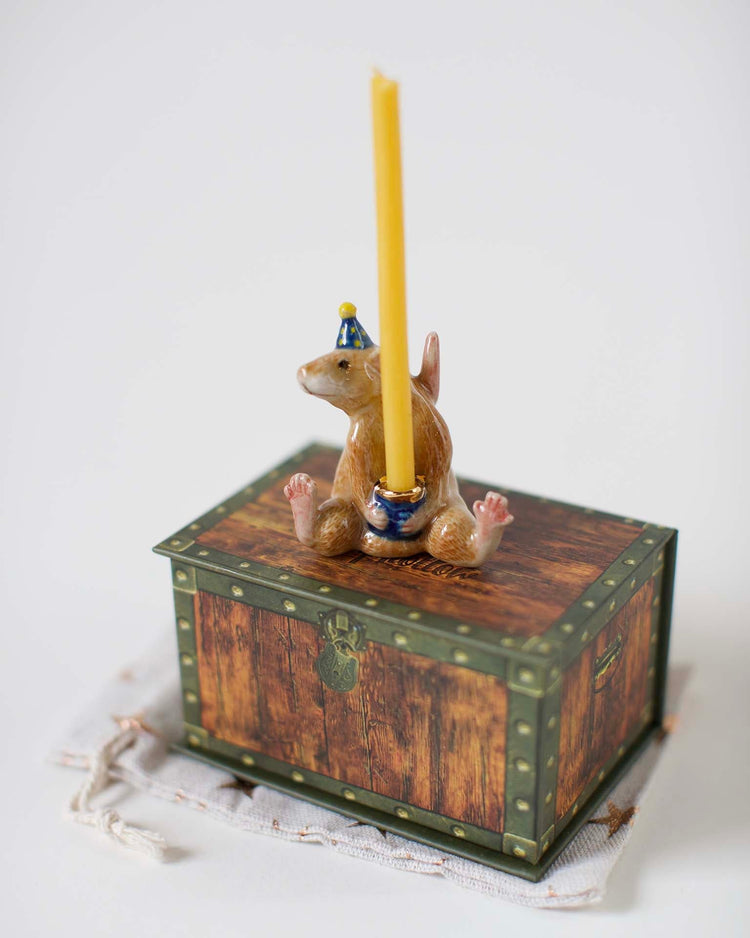 A Camp Hollow hand-painted figurine of a squirrel in a blue hat sitting on a wooden chest, holding a yellow candle upright. The background is plain white.