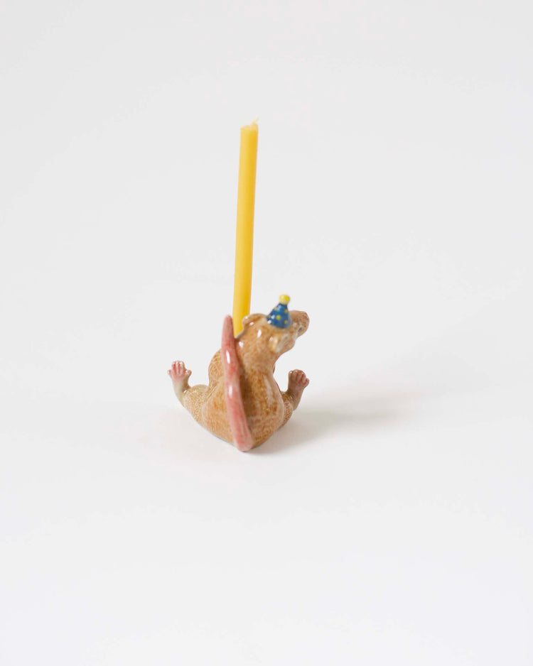 A whimsical, hand-painted candle holder shaped like a small, lying down hippopotamus with a yellow candle inserted on its back.