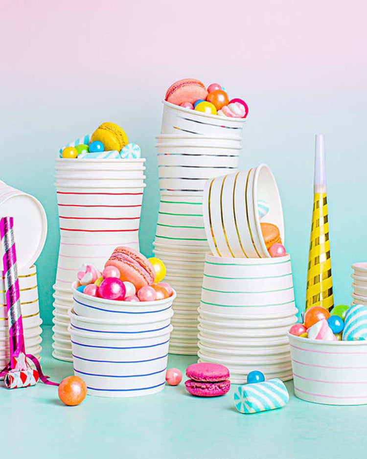 Stacked Silver Frenchie Striped Treat Cups from Daydream Society filled with colorful candies and macarons against a pastel background, with party blowers placed around them.