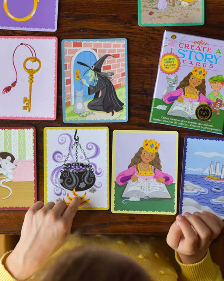 A child's hands point at illustrated story cards featuring various scenes, including a witch with a magic potion, a key, a cauldron, fairy tale characters like a princess reading, a mouse, and a boat. The "fairytale mix-ups create a story" booklet by eeboo is also present, enhancing critical thinking skills.