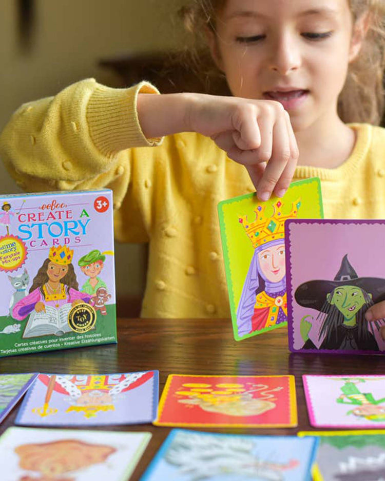 A young girl in a yellow sweater plays with eeboo's "Fairytale Mix-Ups Create a Story" cards, holding a card depicting a queen while various fairy tale characters are spread out in front of her on the table, fostering her critical thinking skills.