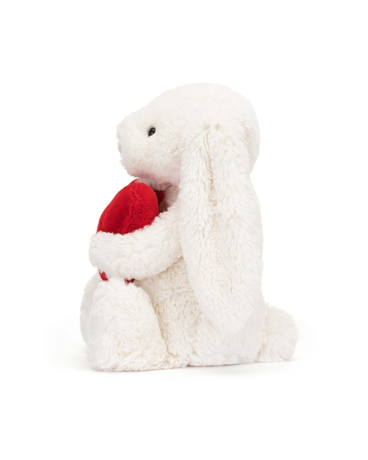 The bashful red love heart bunny little by Jellycat, featuring incredibly soft fur, sits upright holding a red heart-shaped plush, making it an enchanting stuffed animal.