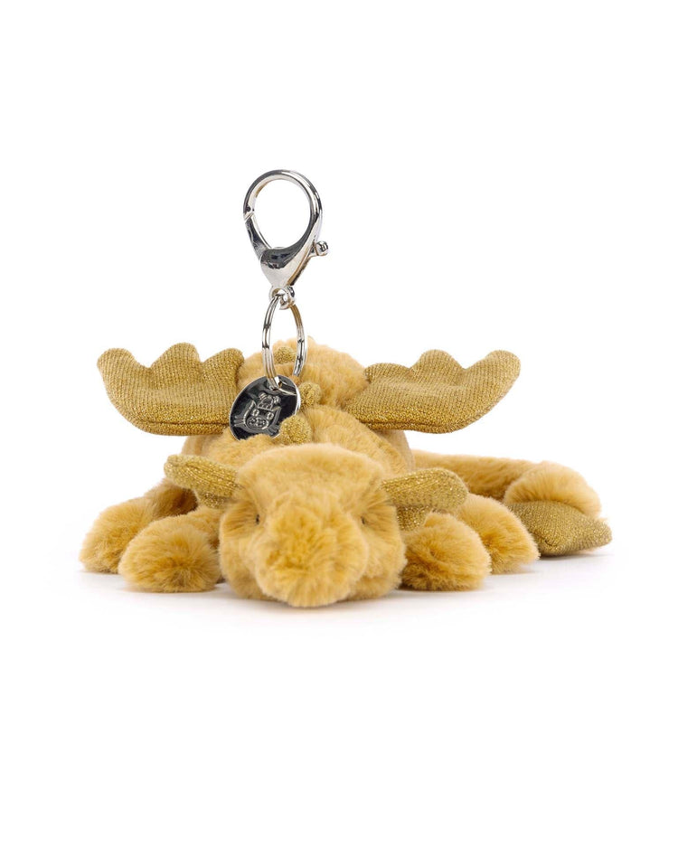 A golden dragon bag charm with intricate detailing and soft texture, lying down with a metallic clasp attached to its head, from the brand Jellycat.