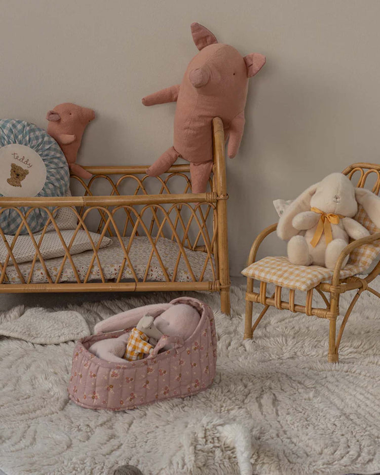 A cozy nursery scene with a rattan crib of stuffed animals, including pigs and a bunny. Small bunny accessories add charm while a baby doll sleeps in the maileg large carry cot in rose on a textured white rug.