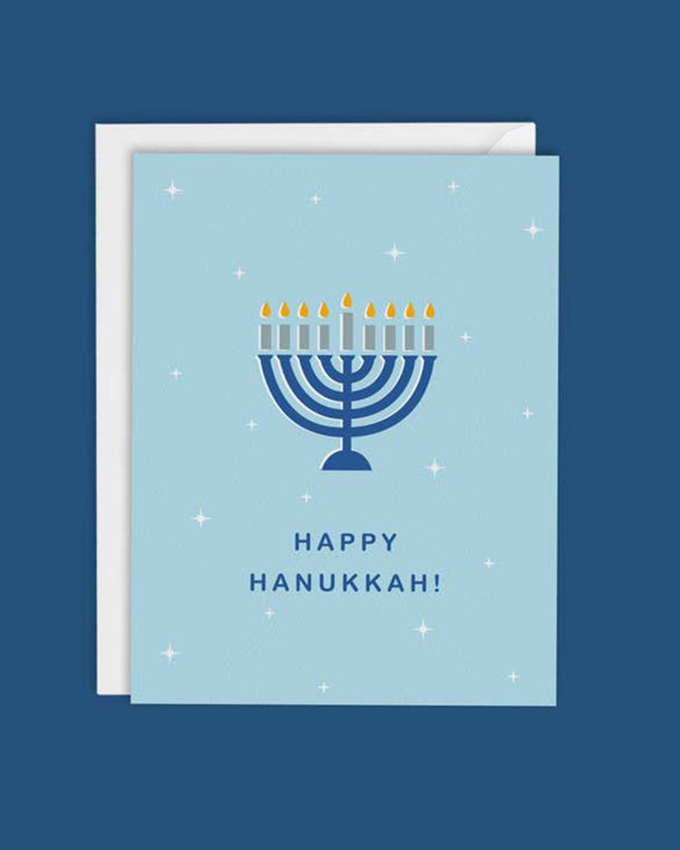 The Happy Hanukkah Menorah Card by MellowWorks showcases a blue menorah with glowing candles, encircled by twinkling stars on a light blue background. The text says "Happy Hanukkah!" and the card is blank inside allowing for your personal message.