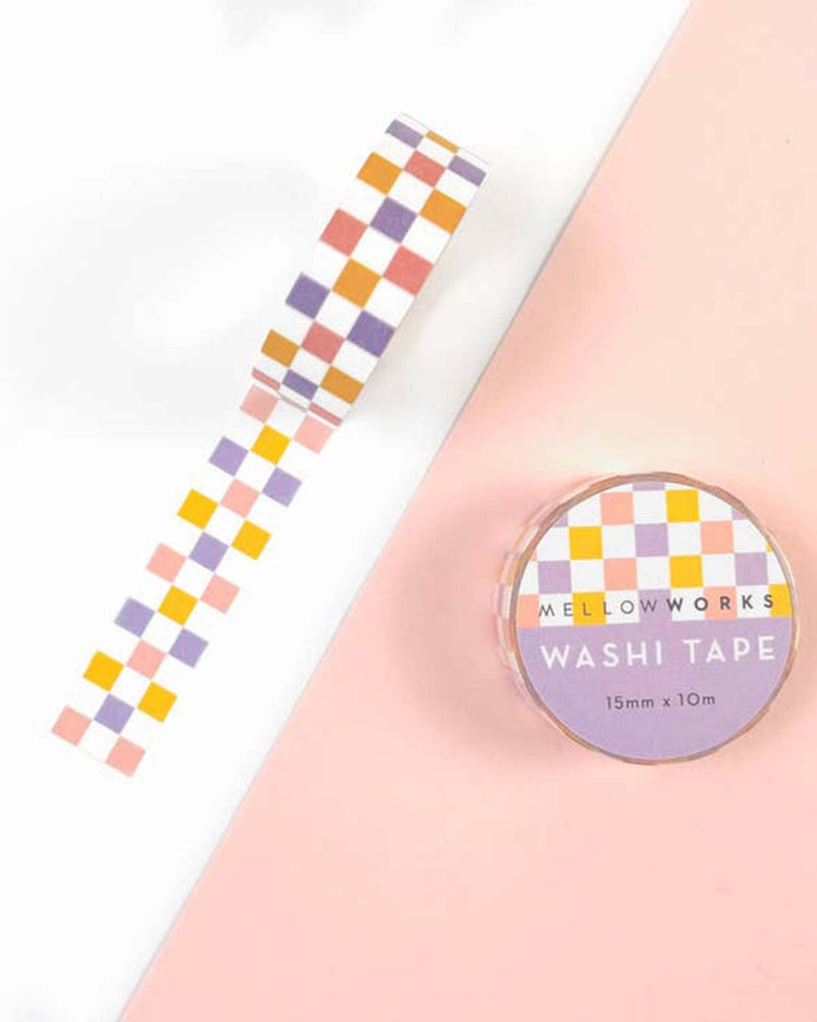 A roll of Mellowworks Lilac Checkerboard Washi Tape in pastel Unicorn colors of orange, yellow, pink, and purple is partially unrolled on a pink and white surface. The label reads "Washi Tape 15mm x 10m." This removable non-residue paper tape is perfect for any project.