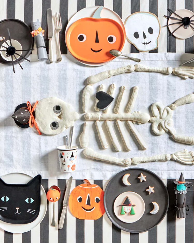 A Halloween table setting with a biscuit-dough skeleton, meri meri reusable jack o’ lantern plates, plus themed cups and cutlery, all on a striped tablecloth.