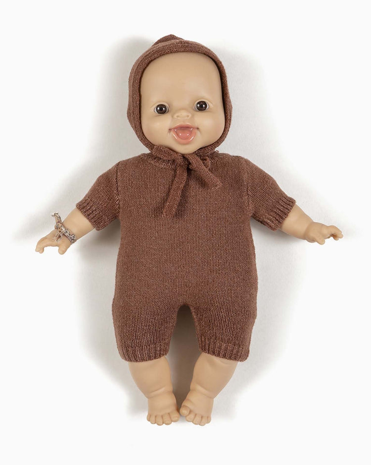 A timeless Minikane Paola Reina baby doll wearing a félix knit jumpsuit in marl caramel and a bonnet, with a bracelet on its left wrist, is lying on a white surface.