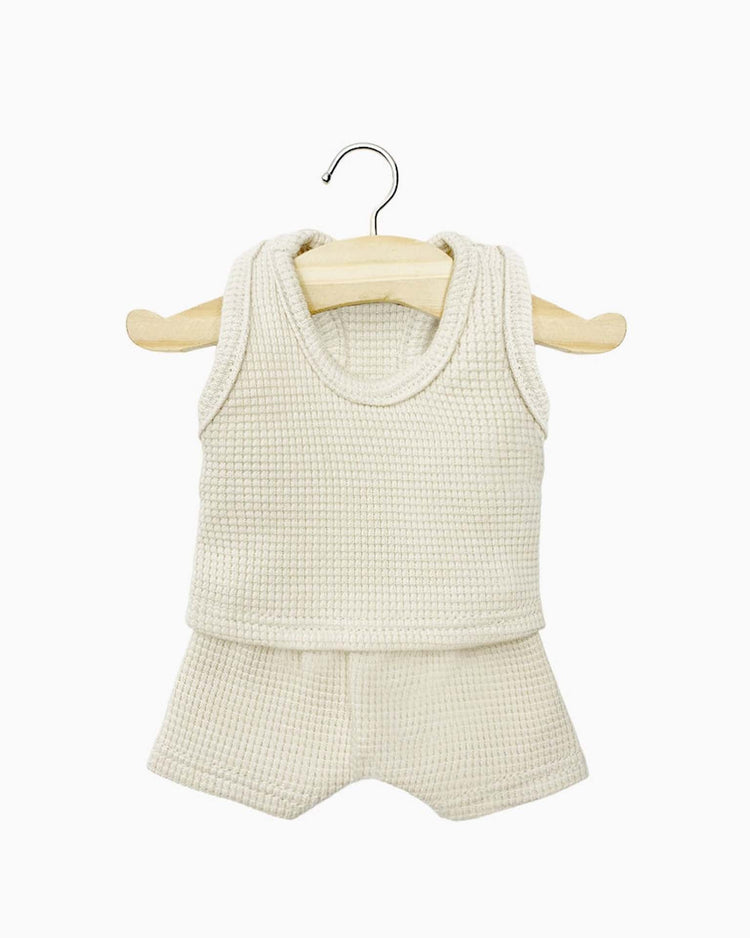 The Les P’tits Basiques waffle tank and short set in linen by Minikane, featuring a cream-colored, sleeveless woven top and matching shorts, is displayed on a wooden hanger against a white background. This set is perfect for Paola Reina Minikane dolls and inspires hours of imaginative play.