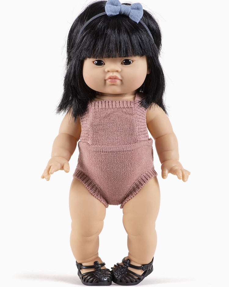 A timeless Minikane doll with black hair, dressed in a Lou knit romper in tea pink, a blue bow headband, and black shoes.