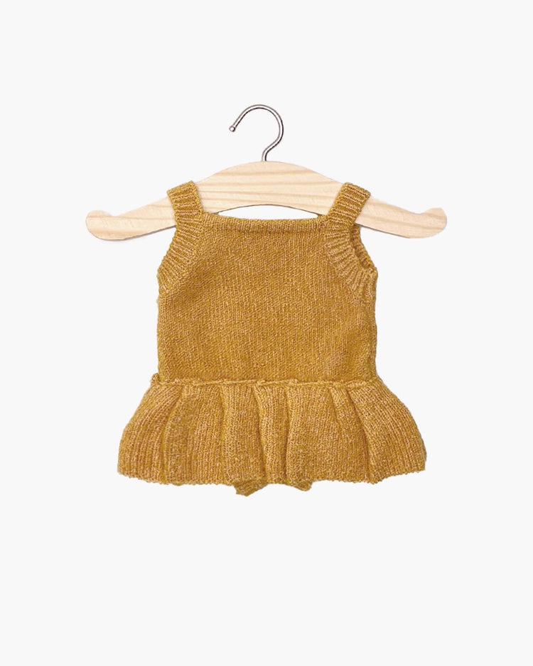 The orléane knit romper in honey, a charming mustard-colored baby outfit with a ruffled skirt by Minikane, hangs on a wooden hanger against a white background.