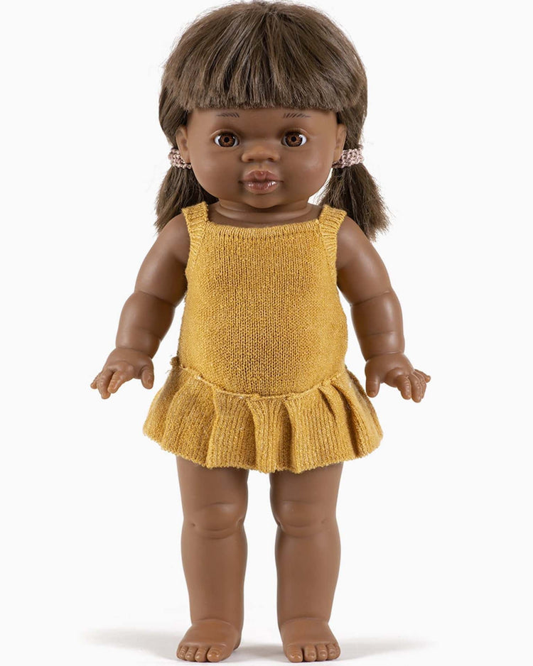 A brown-skinned doll with black hair in pigtails, dressed in an orléane knit romper in honey, stands upright with its arms slightly raised and eyes looking forward. This timeless doll captures the charm of Minikane dolls.