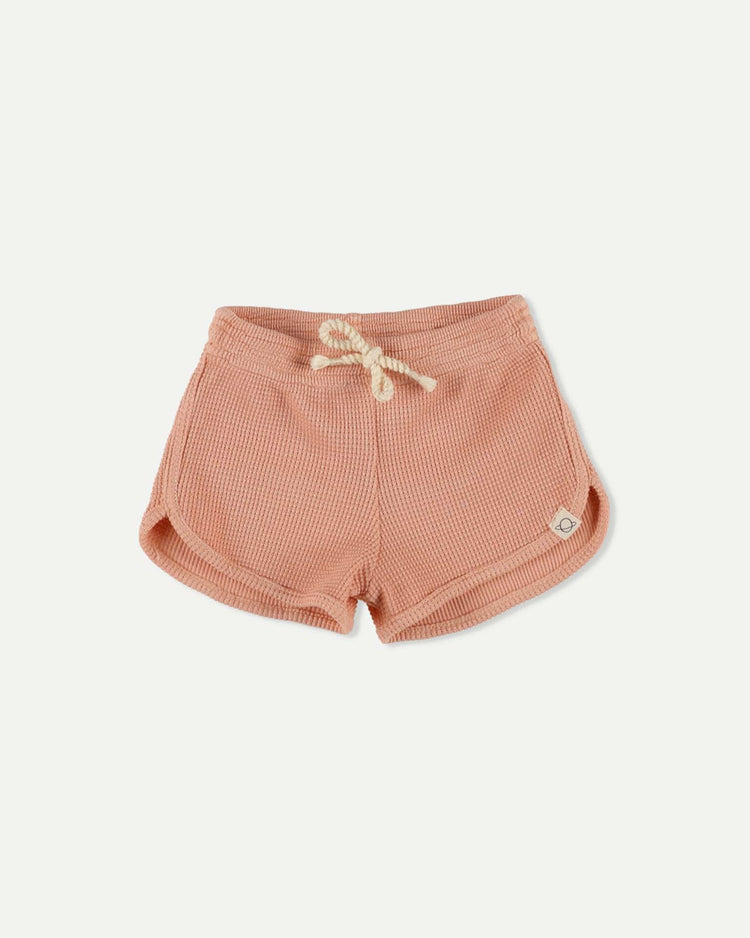 The cozmo nash shorts in pink are organic cotton waffle-knit shorts featuring a cream drawstring, curved hems, and a small round patch on the lower left side, shown laid flat on a white background.