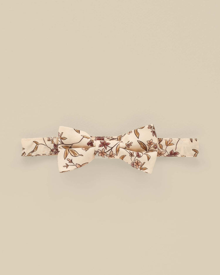 The "bow tie in fig floral" by noralee is crafted from 100% cotton, featuring a charming cream base embellished with a floral pattern of small red and brown flowers, making it perfect for quick and easy wear.