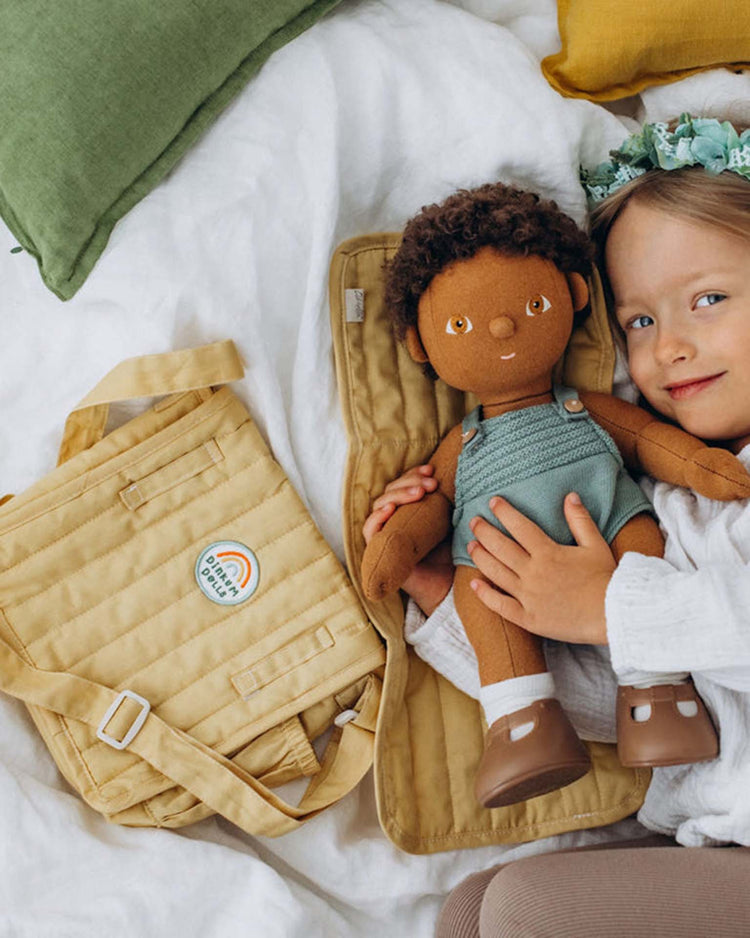 A young child with a flower crown smiles on a bed, holding a brown-skinned doll in green overalls. Next to them is the dinkum dolls carrie convertible changing set in honey, featuring a rainbow patch that reads "dolls over boys.