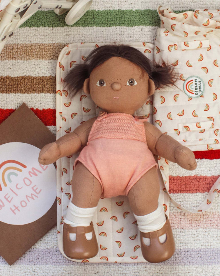 A brown-haired dinkum dolls doll in a pink outfit and brown shoes lies on a striped rug with the carrie convertible changing set in rainbow and other baby accessories around her.