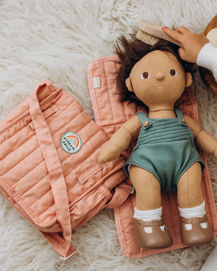 A dinkum dolls carrie convertible changing set in rose features a plush doll with brown hair, green overalls, and brown shoes on a pink quilted mat beside a matching pink changing bag. A hand gently brushes the doll’s hair on a fluffy white rug.