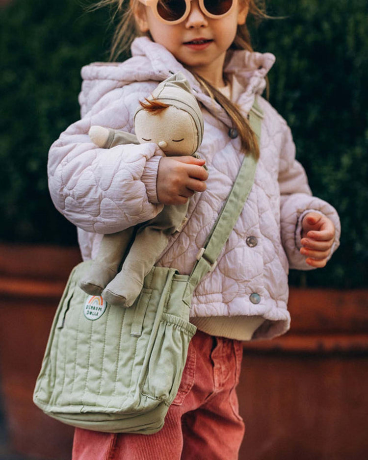 A young child with light brown hair stands outdoors wearing sunglasses and a pale quilted jacket, carrying the Dinkum Dolls carrie convertible changing set in sage—perfect for your little one’s favorite doll accessories.