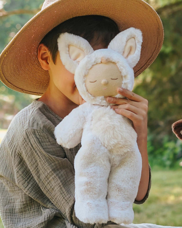 A child in a wide-brimmed hat and grey outfit sits outdoors, holding a dinkum dolls cozy dinkum fox fenix plush that partially covers their face in the sunlight.