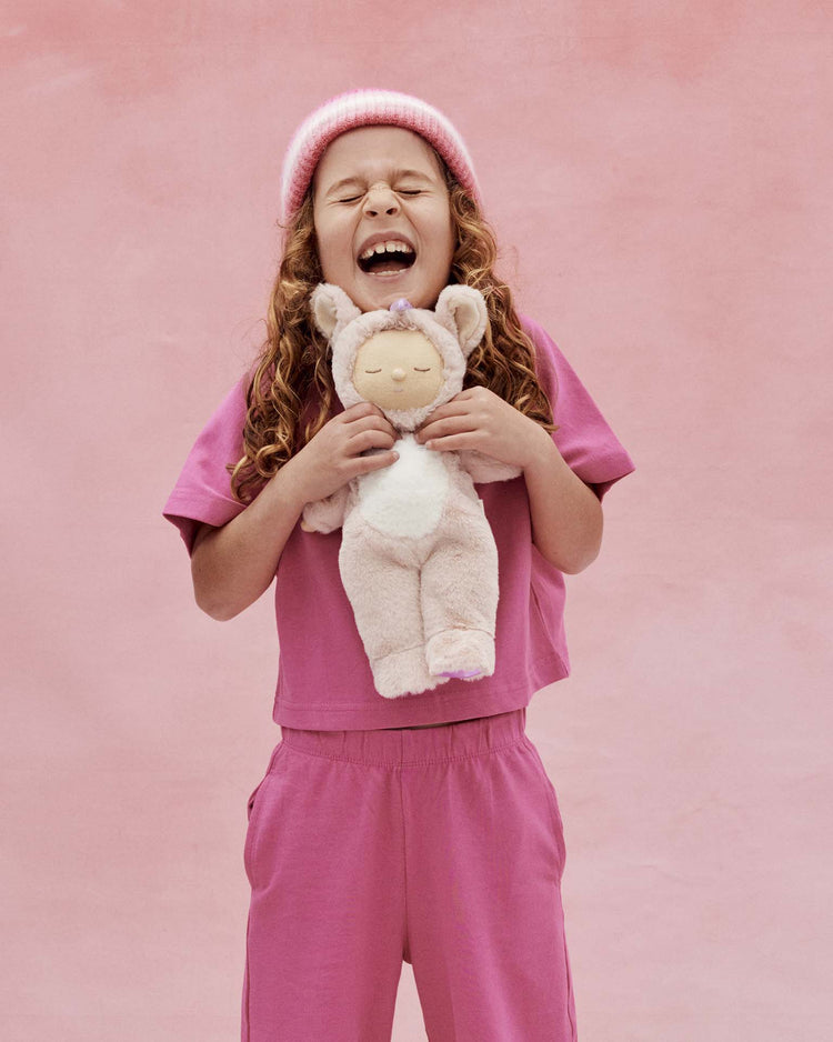 A young child with long curly hair, wearing a pink beanie and outfit, smiles widely against a pink background while holding the dinkum dolls’ cozy dinkum unicorn sugarpie pink toy.