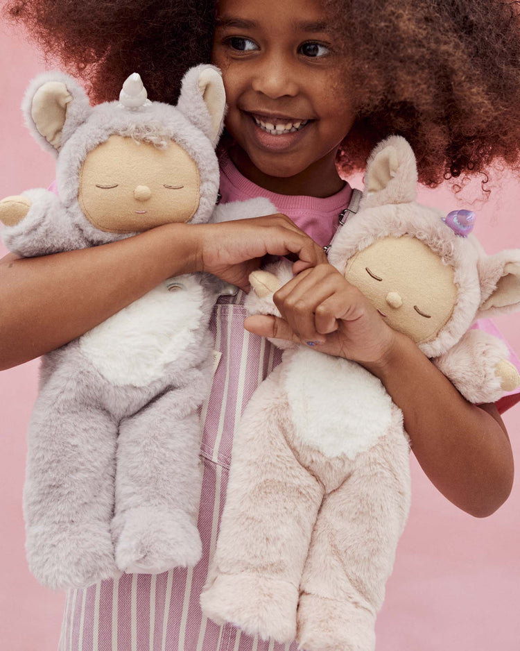 A smiling child with curly hair holds two dinkum dolls cozy dinkum unicorn sweetpea lilac plush toys against a pink background.