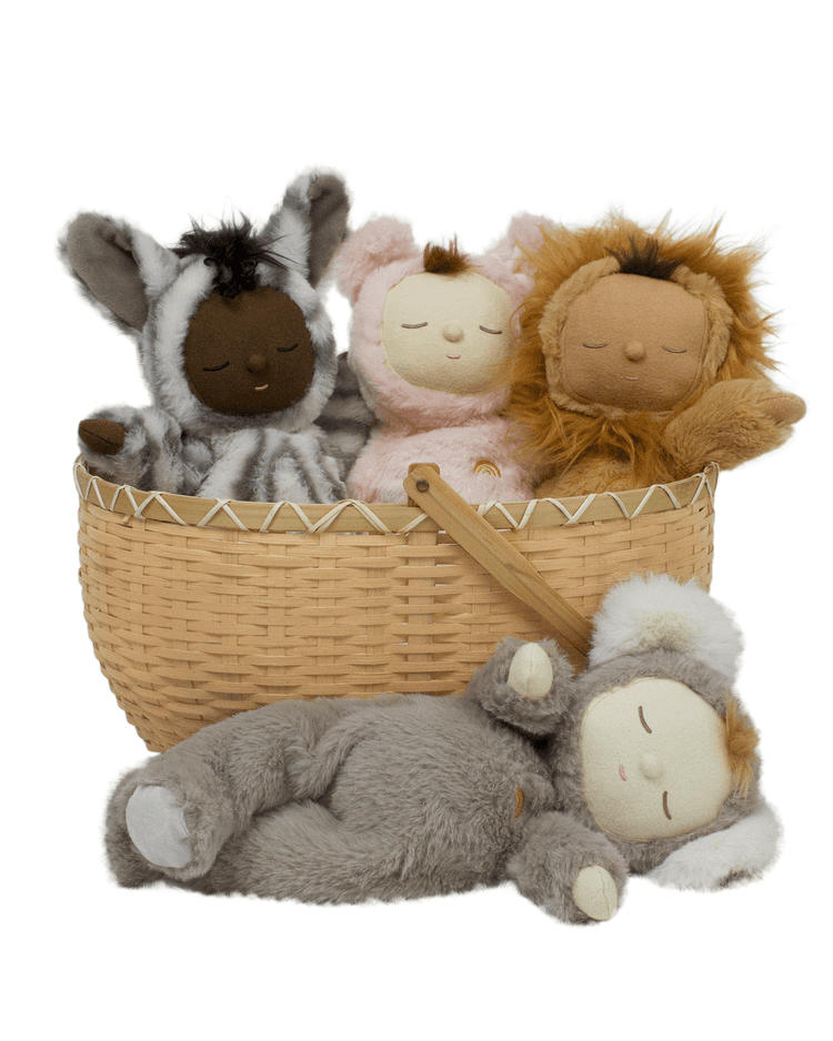 A woven basket holds three plush dolls, including Cozy Dinkum Lion Pip by Dinkum Dolls, plus friends dressed as a pink bunny and the Cozy Dinkum Zebra Mini. A fourth gray plush doll lies in front; all have closed eyes and peaceful expressions.