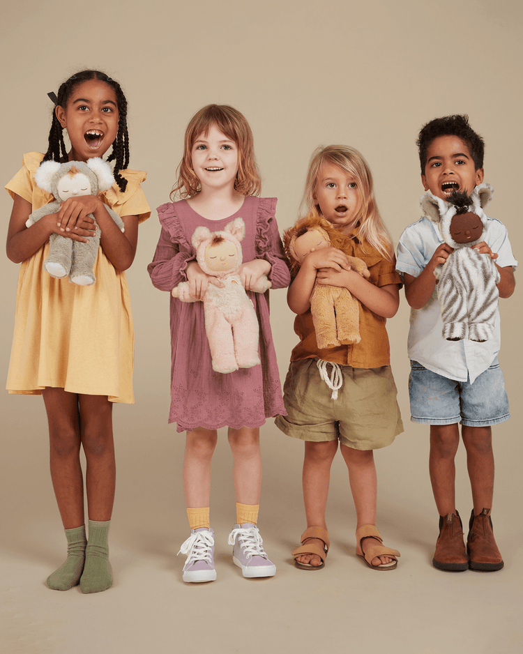 Four young children stand in a row, smiling and holding Cozy Dinkum Zebra Mini plush dolls from Dinkum Dolls. They wear colorful, casual clothes and pose against a neutral beige background.