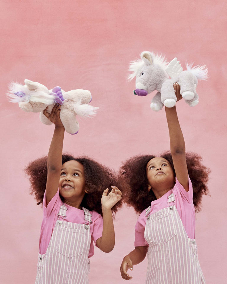 Two young children in matching pink shirts and striped overalls smile as they hold up dinkum dolls' Dinkum Unicorn Glimmer plush unicorn toys against a pink background, looking up at their magical new friends.