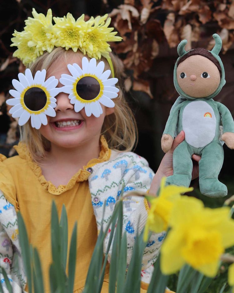 A smiling child in a yellow dress and floral sweater wears daisy sunglasses and a yellow flower headband, holding the dinkum dolls dinky dinkum dolls gideon grasshopper in green—perfect for Backyard Buddies adventures among blooming daffodils.