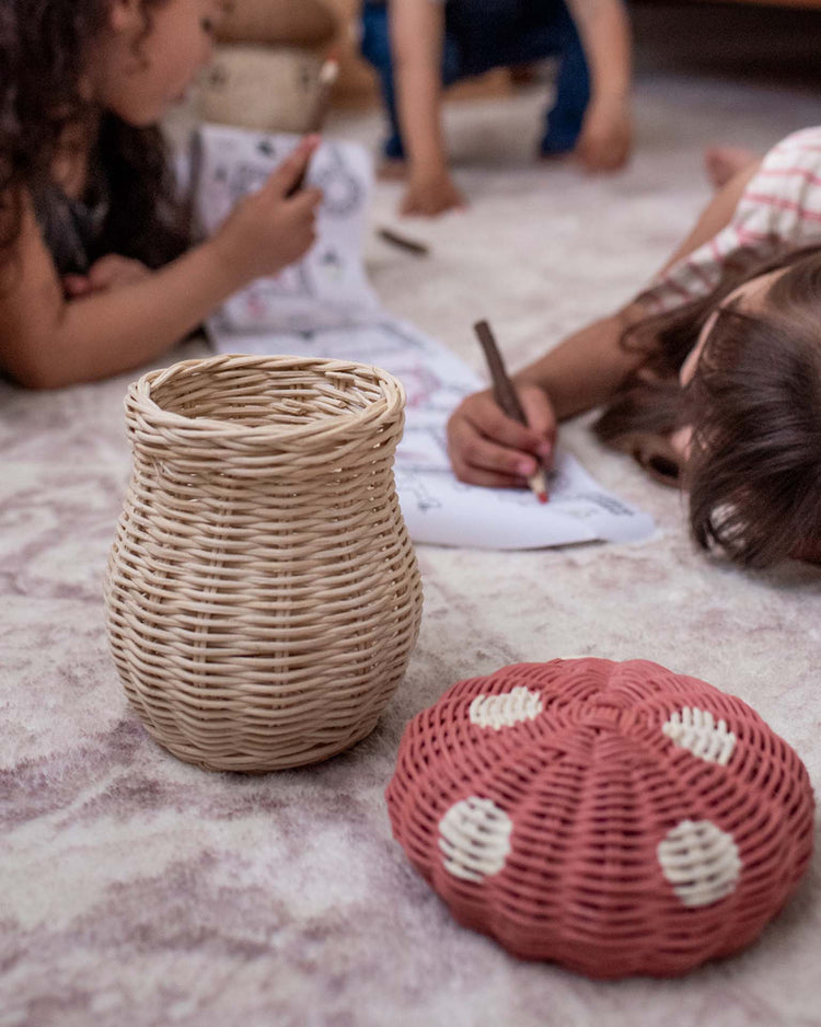A woven basket and the Dinkum Dolls Porcini Basket in Musk with white spots sit on a rug, as children draw and color on paper in the background.