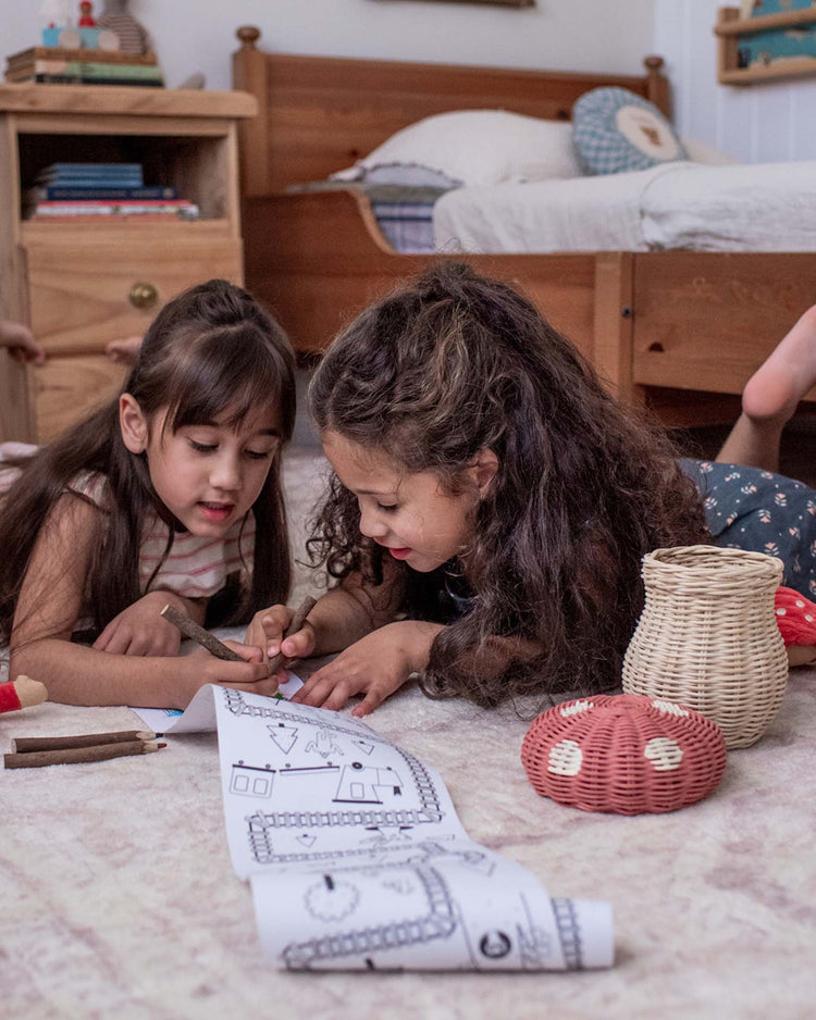 Two young girls color together on the floor, colored pencils and the dinkum dolls porcini basket in musk nearby. The cozy child’s bedroom, with warm wooden furniture, creates a playful atmosphere.
