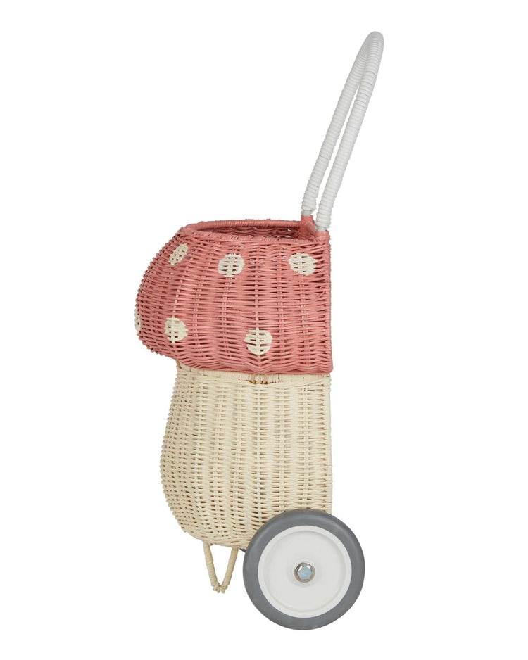 The dinkum dolls rattan mushroom luggy in musk features a cream and pink wheeled design with a white handle and a pink, polka-dotted hood, perfect for whimsical toy storage. Shown upright from the side.