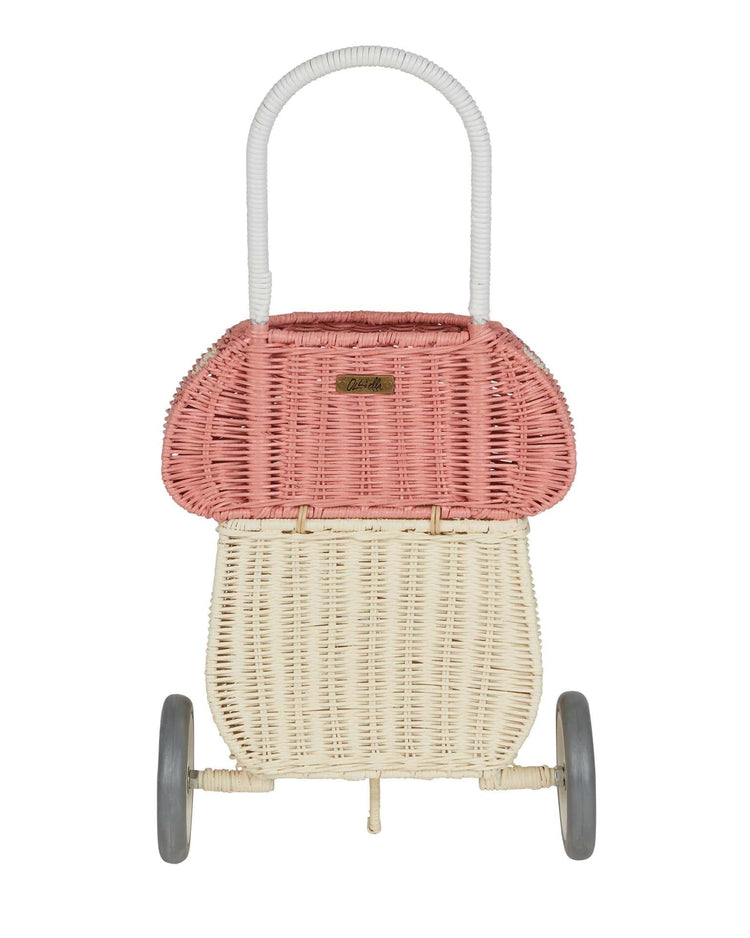 The dinkum dolls rattan mushroom luggy in musk features a cream basket, pink lid, white woven handle, and gray wheels—a charming wheeled stroller for toy storage or play—shown against a white background.