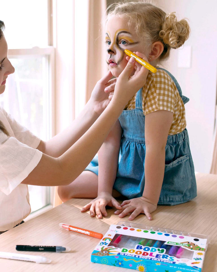An adult is painting a child's face while the child sits on a table. Laid out on the table are art supplies labeled "ooly Body Doodlers Face & Body Crayons Set of 12," which are non-toxic face and body crayons.