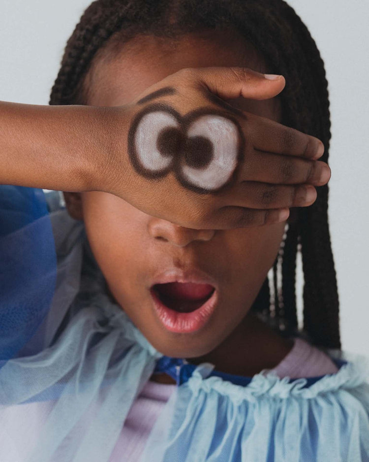 A child with an open mouth covers one eye with a hand decorated using Ooly's Body Doodlers Face & Body Crayons Set of 12, wearing a blue dress.