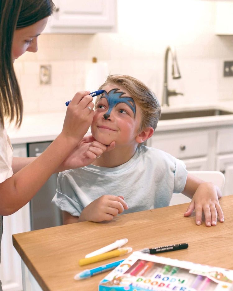 A child sits at a table while an adult applies blue face paint using the "body doodlers face & body crayons" set of 12 by ooly. Various non-toxic markers and temporary body art supplies are spread out on the table.