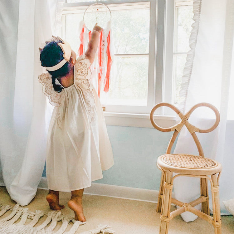 A child in a light dress stands on tiptoes reaching for a dreamcatcher by a bright window, next to the Poppie Bow Chair from poppie toys in a softly lit room.