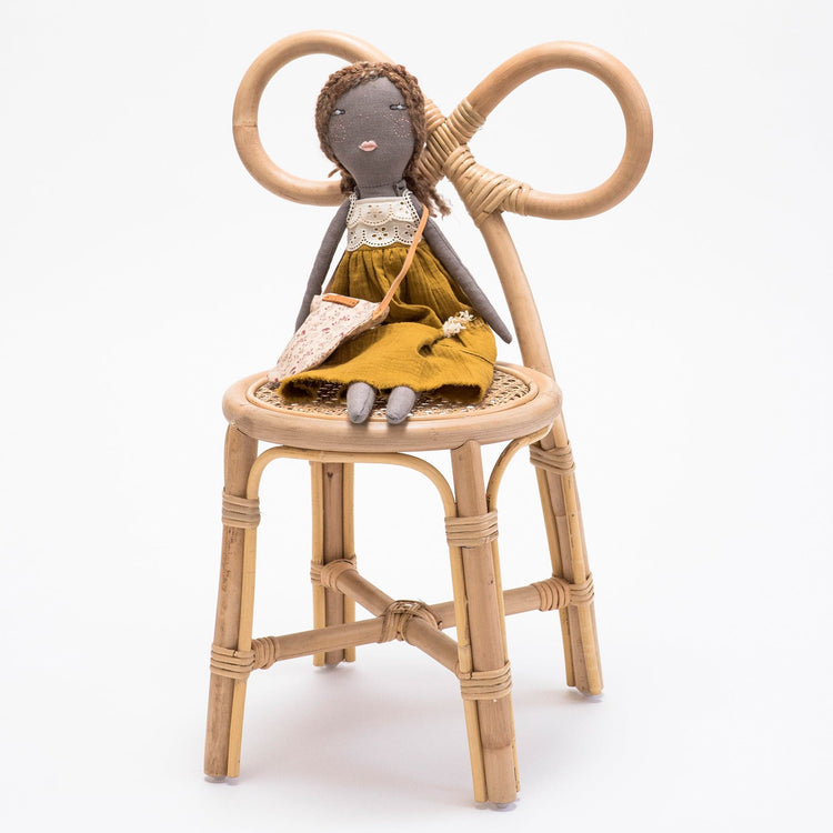 A cloth doll with brown hair, a yellow dress, and a small bag sits on the Poppie Bow Chair by poppie toys against a plain white background.