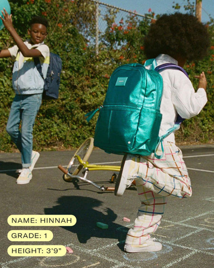 Two children play outside on a sunny day. Hinnah, a first grader who is 39 inches tall, kicks a chalk-drawn hopscotch grid while wearing the STATE Bags Kane backpack in blue checkerboard. Another child walks in the background.