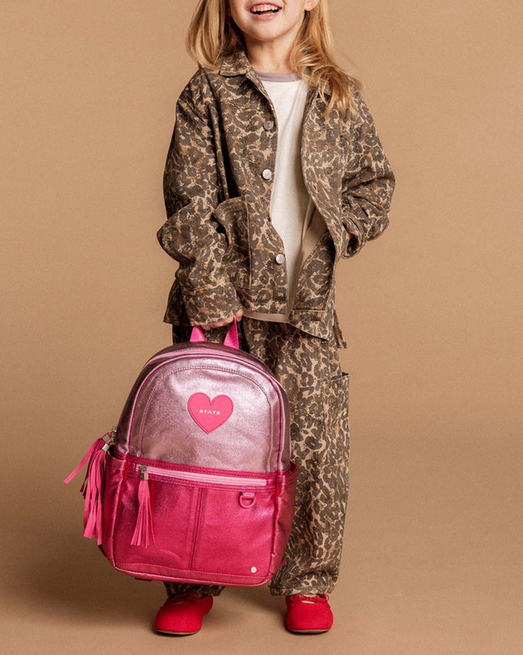 A child in a brown leopard-print outfit and red shoes holds the STATE Bags Kane Backpack in Pink + Hot Pink Colorblock, which features a heart-shaped patch, against a plain tan background.