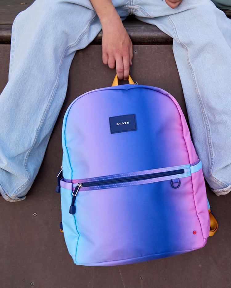 A person in light jeans sits, holding the STATE Bags Kane Large Double Pocket Backpack in a blue and pink gradient, featuring a front zipper pocket and the brand’s black label. Their hand rests on top of the backpack.