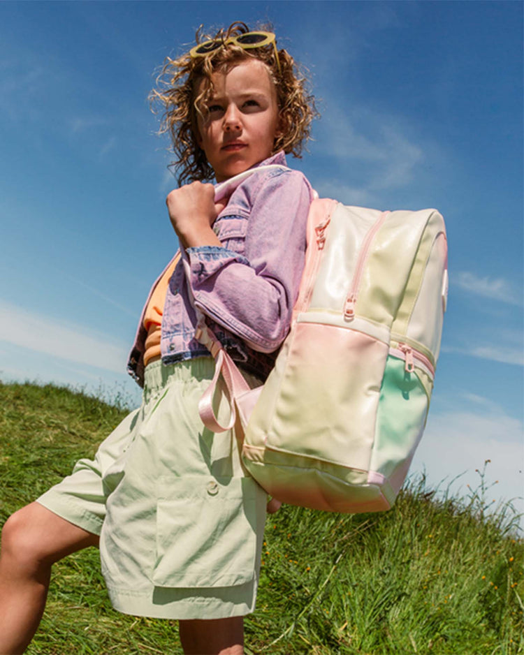 A young person with curly hair stands outdoors in pastel-colored clothing, carrying the STATE Bags Kane Large Double Pocket Backpack in Tie Dye Patchwork over one shoulder, with grass and a blue sky behind them.