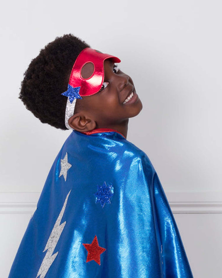 A smiling child wears a vibrant red superhero mask and a blue cape adorned with stars and a lightning bolt, posing against a simple white background—showcasing the perfect look from our meri meri blue superhero costume, an ideal set for any kids’ superhero dress-up.