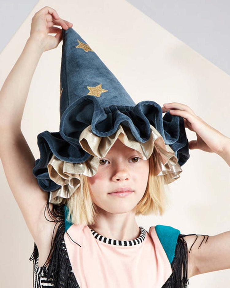 A child wearing the meri meri pointed blue hat, which features gold stars and ruffled edges, gently touches the Halloween accessory with one hand while looking forward.