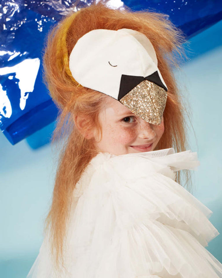 A child with red hair wears a white ruffled costume and a bird mask with a glittery beak, smiling over their shoulder against a blue and light blue background. Their playful look is completed by delicate tulle wings from the meri meri swan cape dress up set, adding an extra touch of whimsy to the scene.