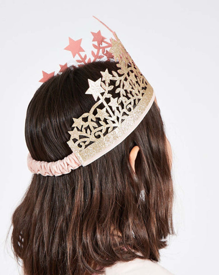A person with shoulder-length brown hair is seen from behind, wearing the meri meri star crown, a gold and pink star-patterned dress-up headband, against a plain white background.