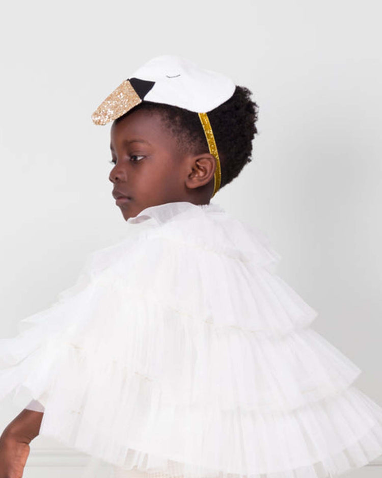 A child wearing the meri meri swan cape dress up set—featuring a layered white ruffled cape and a swan-themed headband with a gold beak—stands sideways against a plain light background, beautifully capturing the essence of swan-inspired play. Delicate tulle wings complete this whimsical look, making it perfect for imaginative dress-up fun.