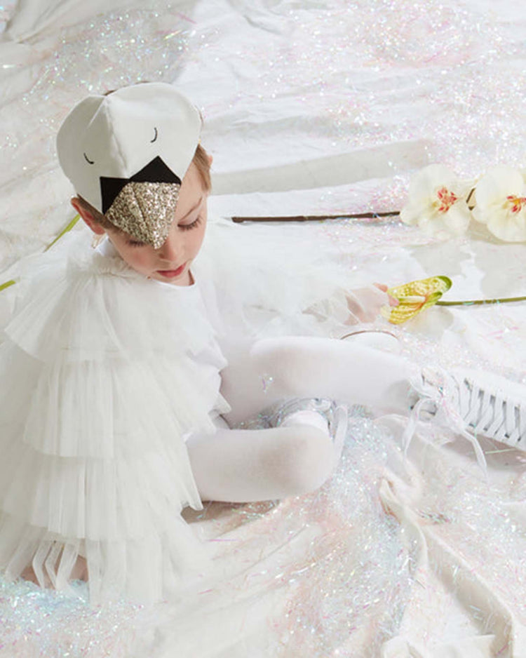 A child wearing the Meri Meri Swan Cape Dress Up Set, featuring a white swan costume with a sparkly gold beak and tutu, sits on a shimmering iridescent surface beside white flowers and a butterfly wand, gazing downward. Delicate tulle wings softly flutter behind them, while a plush velvet headpiece completes the enchanting ensemble.