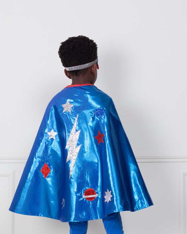A child wearing a shimmering blue superhero cape adorned with stars, a lightning bolt, and planets stands with their back to the camera. Part of the meri meri blue superhero costume, the look is finished with a silver headband—perfect for kids' dress-up adventures.